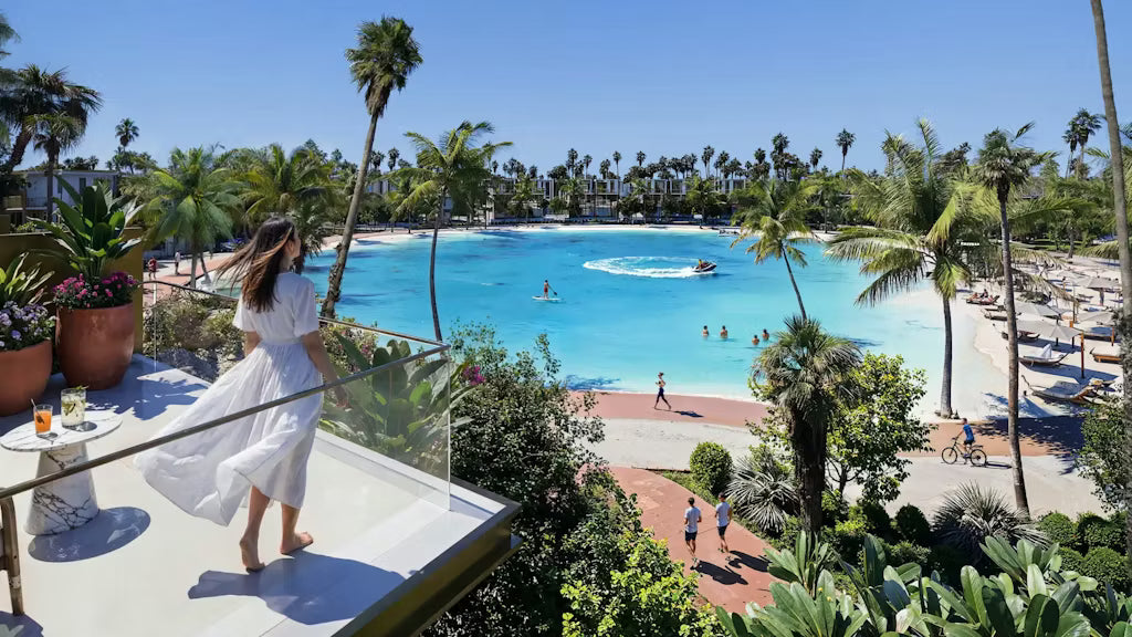 Woman in a white dress standing on a balcony overlooking a pool with palm trees and people in the water.